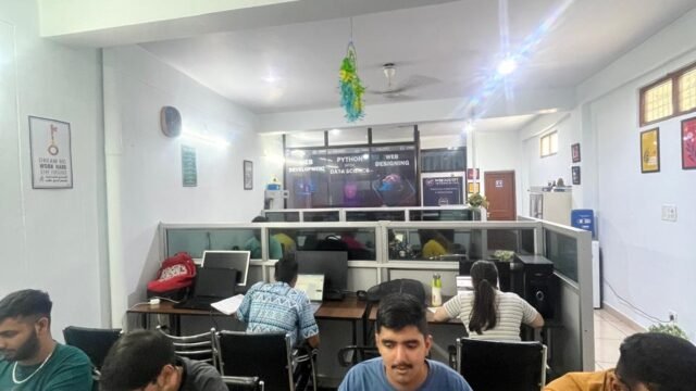 Open office coworking space with several coworkers at desks and laptops, glass partition walls in the background, bright lighting.