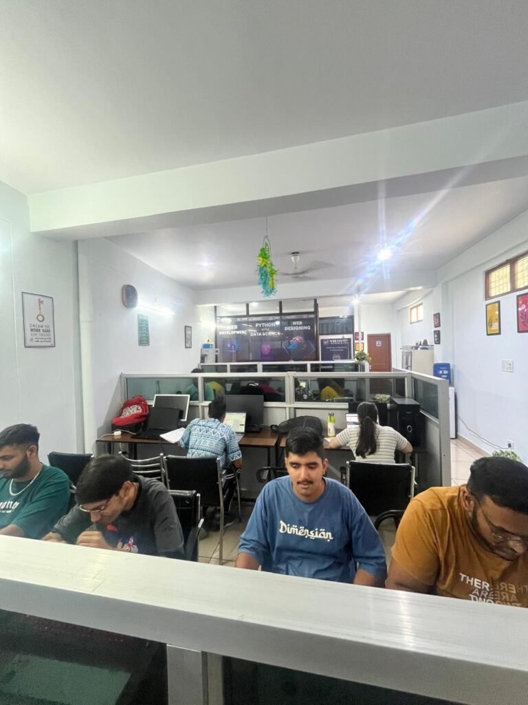 Open office coworking space with several coworkers at desks and laptops, glass partition walls in the background, bright lighting.